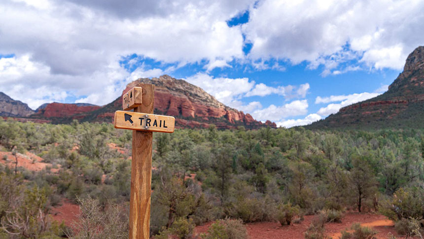 A trail signpost out in the Arizona Desert with mountains and a blue sky with clouds in the background.